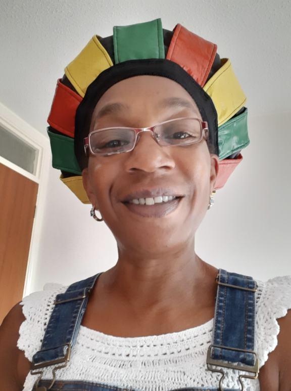 A close-up portrait photo of a black woman smiling, looking at the camera. She wears glasses, small hoop earrings, a white crochet top, denim dungarees and a black leather hat with yellow, green and red leather stripes. There is a white wall behind her and a wooden door.