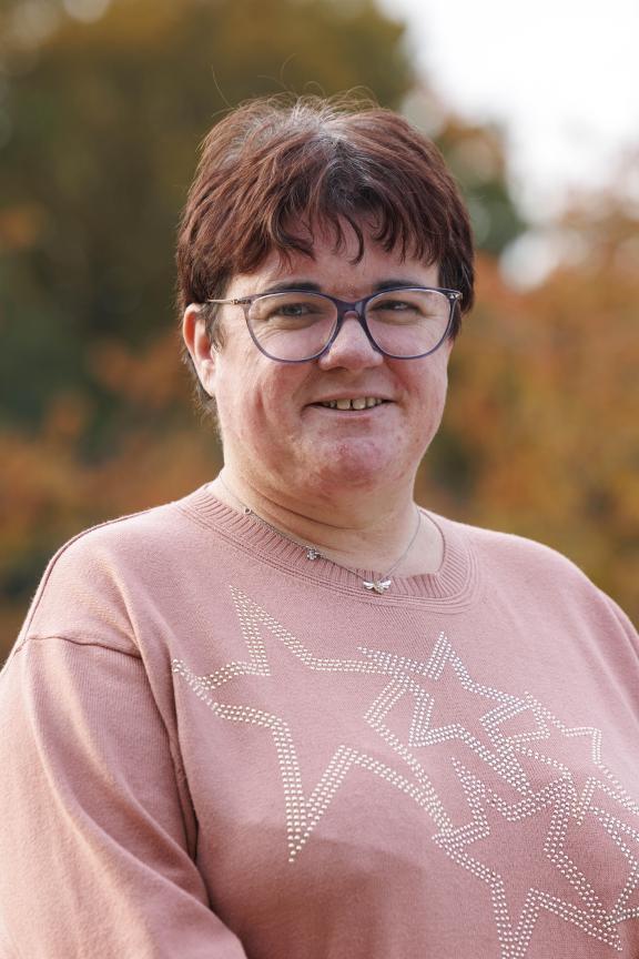 A woman smiling with cropped reddish brown hair wearing glasses and a pink jumper with stars. A blurred background indicates an autumnal outdoor setting with yellow and green leafed trees.
