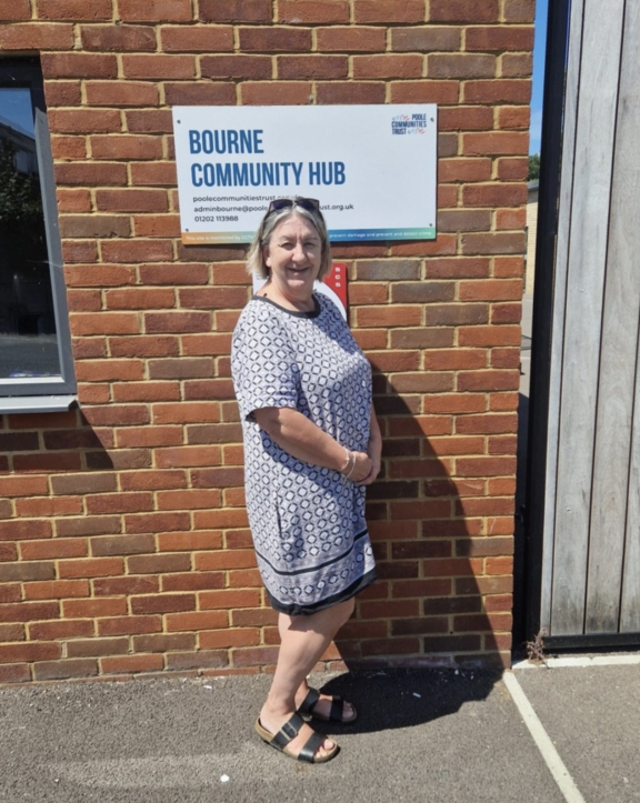 An older woman wearing sunglasses on her head wears a blue and white patterned knee-long dress and black sandals. She faces the camera and has her hands clasped, standing against a brick wall with a sign on it which reads "Bourne Community Hub".
