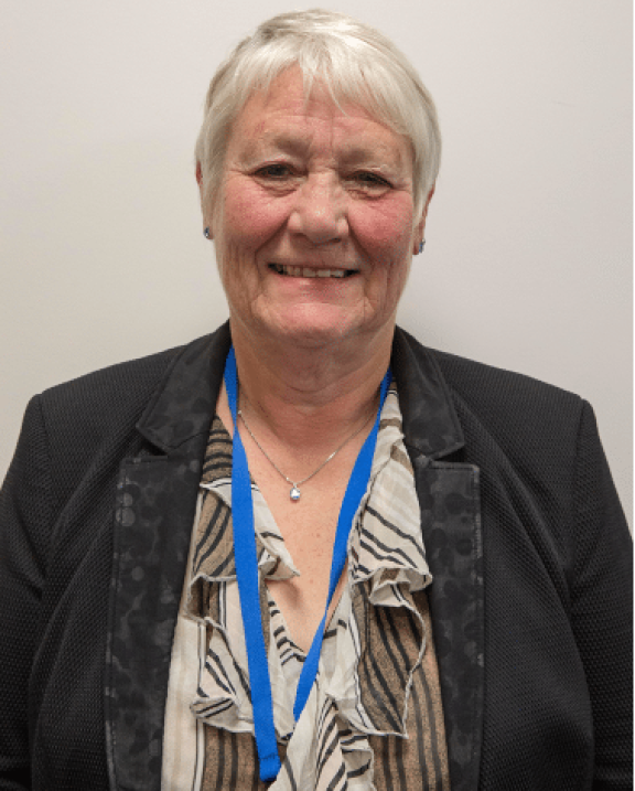 A head and shoulders portrait of older woman with white short hair smiles at the camera. She wears small earrings, a pendant necklace, a blue lanyard, a stripey beige and brown striped ruffle top, and a black suit jacket against a plain background.