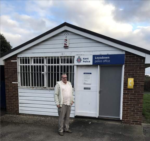 A man with grey hair, sunglasses, a white shirt, grey hoodie, taupe trousers and shoes stands outside a small brick and shiplap building. Over the front door there are two signs - Kent Police and Leysdown police office. Blue sky with white and grey clouds can be seen in the background above the police station building.