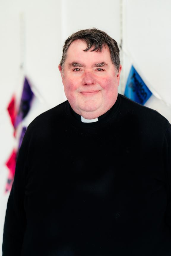 A smiling man in clerical attire, wearing a black shirt with a white clerical collar. He has brown hair, is clean-shaven. Behind him are several flags.