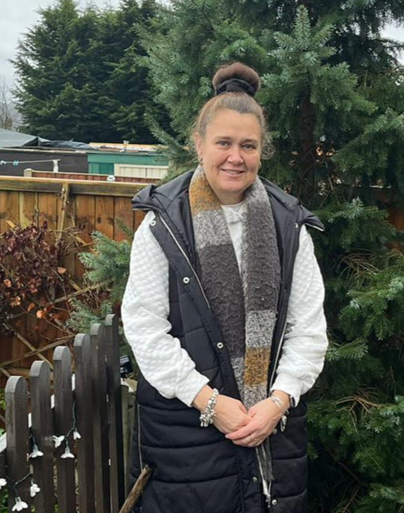 A woman with her hair tied up stands in a garden, smiling at the camera. She is wearing a white sweater, a black vest, and a striped scarf. In the background, there is a wooden fence and a large evergreen tree.