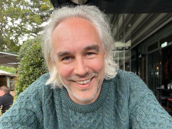 Smiling man with shoulder-length gray hair and a beard, wearing a green cable-knit sweater, sitting at an outdoor café with greenery and string lights in the background.