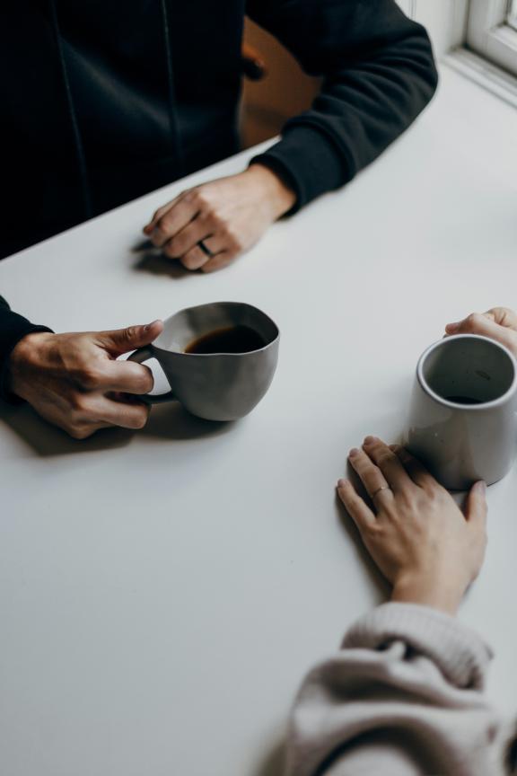 A close-up image of two people's hands sitting opposite from each other at a white table by a window. They are both holding grey mugs.