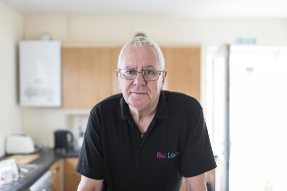 A man with grey hair and glasses wearing a black polo neck shirt with 'Big Local' text embroidered on it. He is standing in a kitchen, leaning against the work surface as he looks into the camera lens.