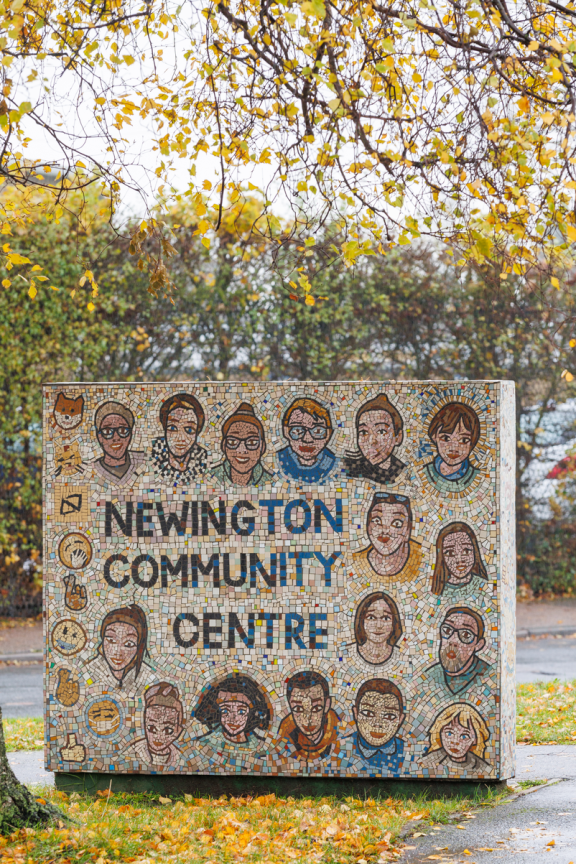 A large square colourful mosaic block depicts different adults and children and emojis surrounding the words "Newington Community Centre". It is positioned under tree branches with yellow leaves, along a pavement and road. There are more trees and bushes in the background and a white sky.