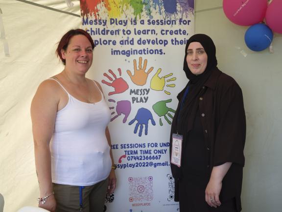 Two women standing together in front of a colourful promotional banner for 'Messy Play' sessions. The banner features rainbow-colored handprints arranged in a circle around the text 'MESSY PLAY' and explains that these are sessions for children to learn, create, explore and develop their imaginations. The woman on the left is wearing a white tank top and has reddish-brown hair, smiling at the camera. The woman on the right is wearing a black hijab and dark clothing with what appears to be an ID badge. Pink and blue balloons are visible in the background.