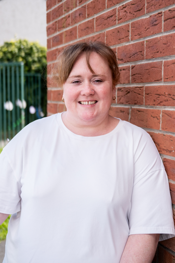 A young woman with her hair up wears a white tshirt and small gold hoop earrings while smiling at the camera. She is leaning against a red brick wall. A green fence and bush can be seen in the background.