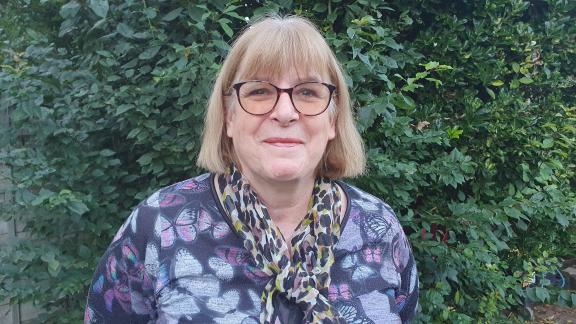 A middle-aged woman with shoulder-length blonde hair and a fringe stands smiling in front of green bushes. She wears dark-rimmed glasses and is dressed in a purple top with a colorful butterfly pattern . Around her neck is a leopard print scarf.
