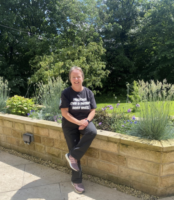 A middle-aged woman with short hair smiles at the camera while perching on a brick wall of a raised beds planted with flowers and grasses. She is wearing purple trainers, black trousers and a black tshirt. There are mature trees behind her and a green lawn.