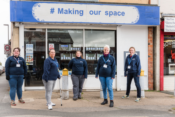 Five Selby Big Local partnership members stand outside their community hub. They are wearing navy blue fleece tops and blue lanyards. The sign above the hub reads '# Making our space'.