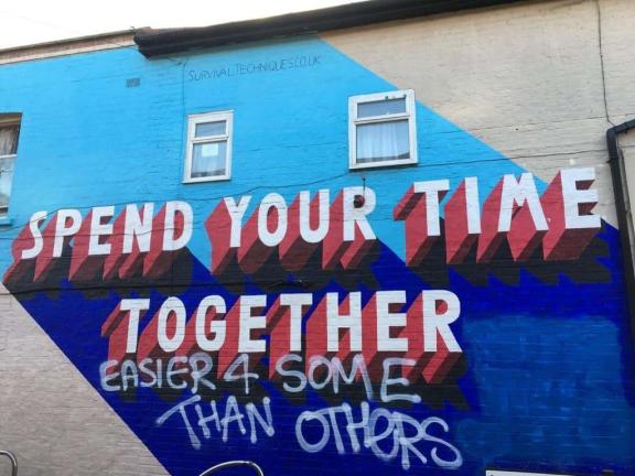 A mural with half light-blue background and half bright blue painted in diagonal. On top of that reads ’Spend Your Time Together’ in 3D style white letters with red shadows. Graffiti below the mural reads 'Easier for some than others" in white spray paint.