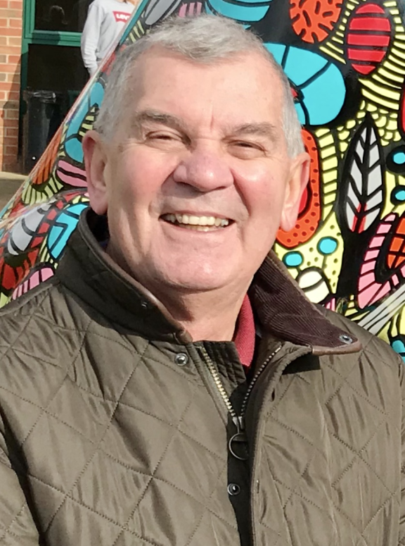 A smiling older man with gray hair wearing a khaki quilted jacket over a coral polo neck shirt stands in front of a vibrant street art mural. The colourful artwork behind him features bold patterns in blues, oranges, reds, and yellows with geometric shapes, floral motifs, and abstract designs. The man appears cheerful and relaxed, positioned against this dynamic urban art backdrop.