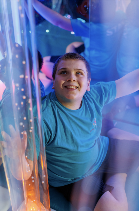 A young boy with short brown hair wearing a turquoise/teal t-shirt smiles broadly while playing in an interactive light installation or sensory play area. He is surrounded by translucent tubes or columns that are illuminated with colorful lights - primarily blue, purple, and orange/amber hues. The lighting creates a magical, immersive environment with glowing effects and sparkles of light throughout the space.