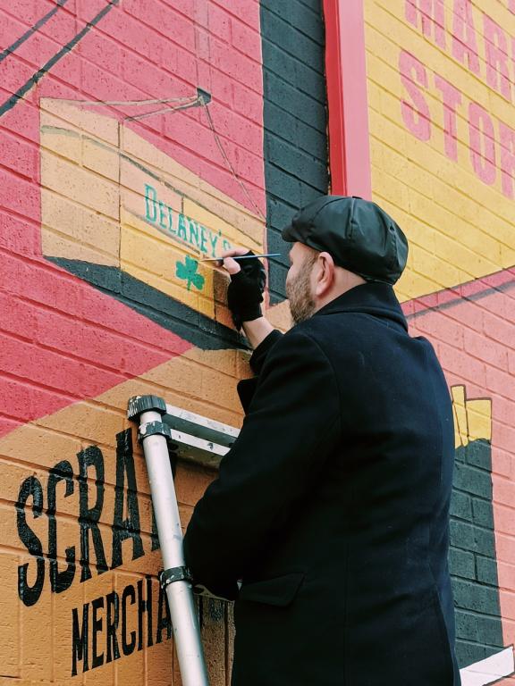 A man wearing a black coat and flat cap stands on a ladder painting details onto a colourful mural on a brick wall. The mural includes an illustration of a wooden crate labelled “Delaney’s” with a small green shamrock, along with large painted lettering on the wall.
