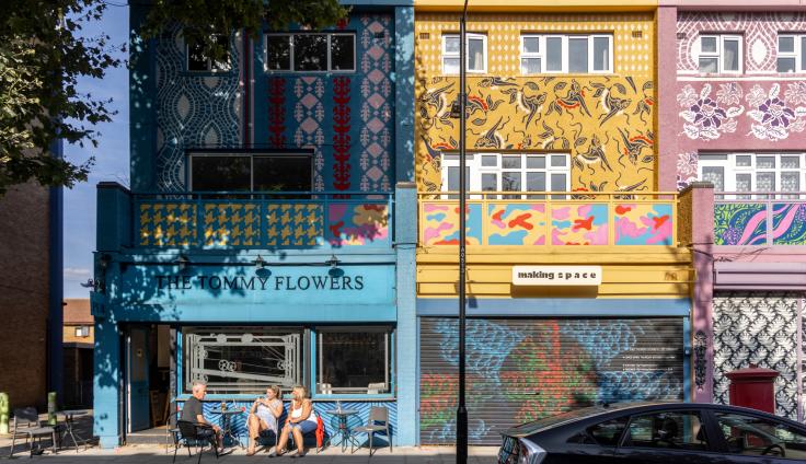 A bright shop-front in teal colours in a row of shops, with a sign above reading 'The Tommy Flowers' and people sitting in chairs on the pavement.