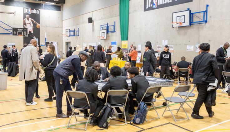Groups of people standing and talking, and students in uniform sitting at a table inside a gym