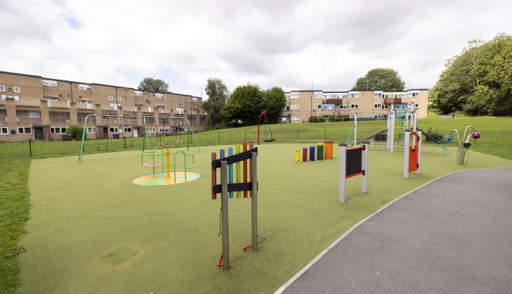 Green play park area with a tarmacked path, swings, roundabout and colourful play equipment