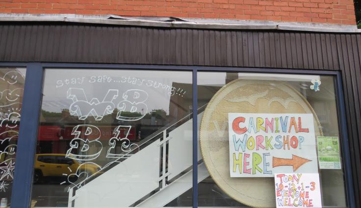 A colourful handmade sign inside of a window, that reads "Carnival workshop here, today 10-12 and 1-3, everyone welcome".