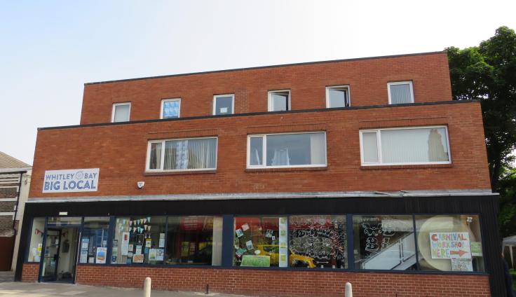 A three-storey red brick building with glass frontage on the ground floor, containing art, bunting and community notices. Above the open double-doors is a white sign with blue text, reading "Whitley Bay Big Local".