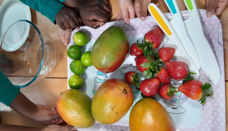 Eight hands reach towards a tray with a variety of fruit on it including strawberries, mangoes and limes.