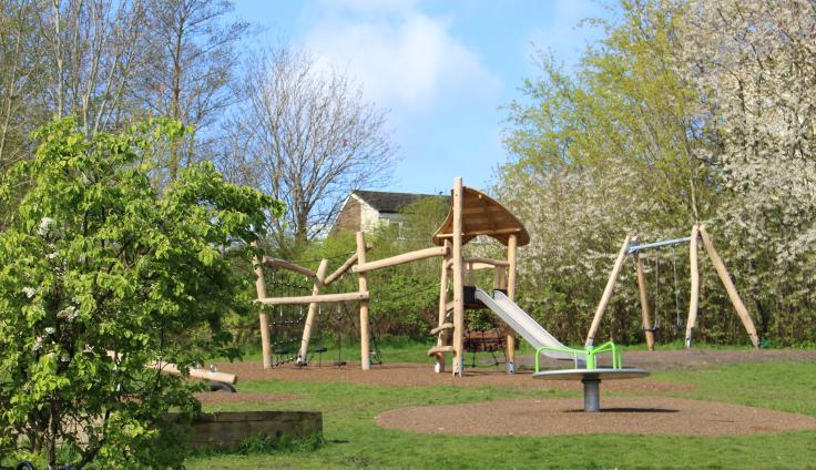 A green, sunny play park with wooden climbing equipment and a slide.