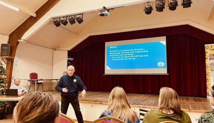 A man wearing a black shirt and trousers stands animated in front of a screen with a blue slide on it, set against a red curtain on a stage. Five people watch him present.