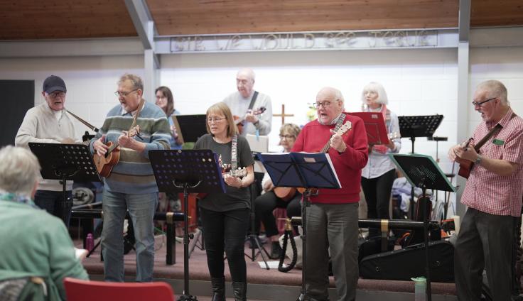 A group of older men and women play instruments and sing in front of music stands.