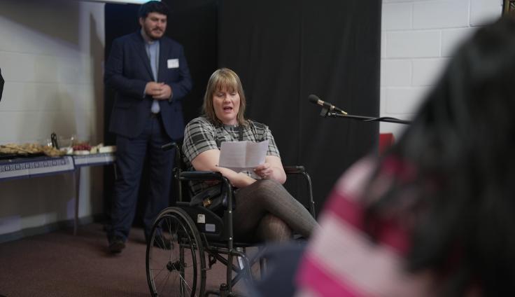 A woman sits cross-legged in a wheelchair as she gives a speech into a microphone in a community hall. Blurred behind her stands a priest.