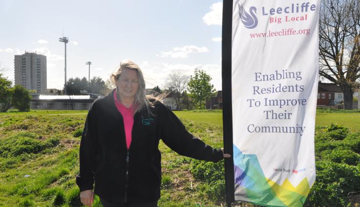 A woman with blonde hair, a pink polo shirt and a black fleece stands next to a flag that reads "Leecliffe Big Local. www.leecliffe.org Enabling residents to improve their community". She stands in a green field, with trees, houses and a tower block in the background.