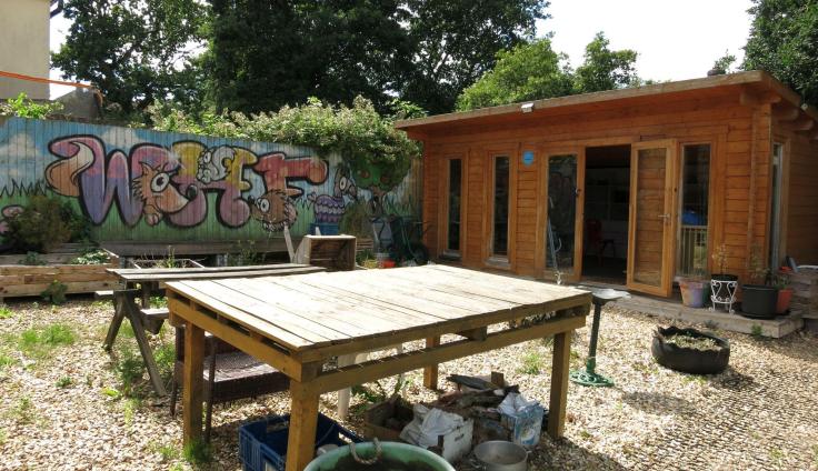 A wooden table in the sunshine in front of a wooden building, with a colourful spray-painted mural on a wall in the background.