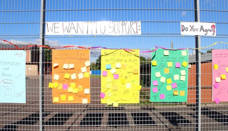 Brightly-coloured posters covered in post-in notes, attached to a steel mesh fence.
