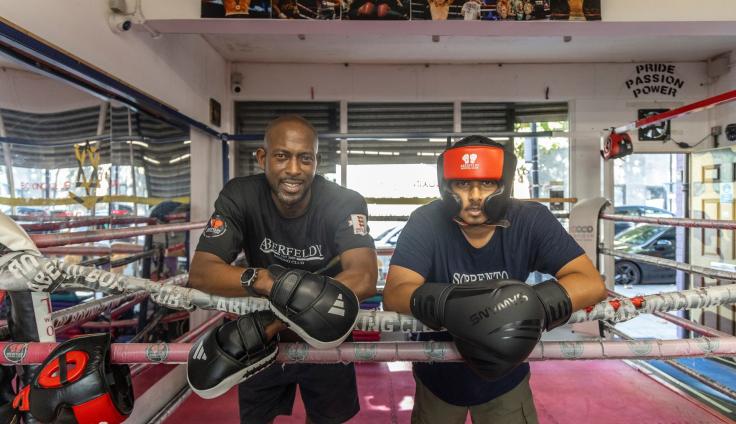 Two men wearing boxing gloves leaning on a boxing ring rope and smiling at the camera.