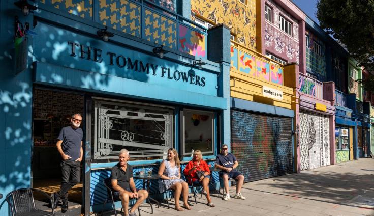 A bright shop-front in teal colours in a row of shops, with a sign above reading 'The Tommy Flowers' and people sitting in chairs on the pavement.