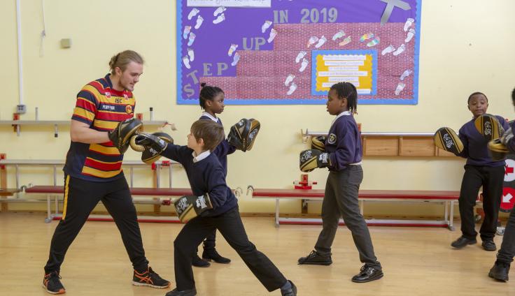 A group of children in school uniforms sparring with a boxing instructor in a striped top.