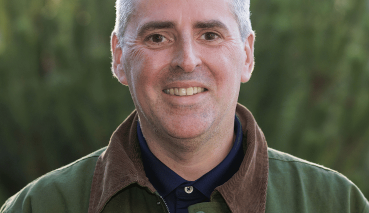 A man with grey hair and brown eyes wearing a green jacket with a brown collar and a navy shirt smiles at the camera. He stands against a blurred background of green bushes.
