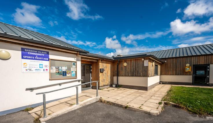 One-storey white building with wooden panelling and a blue sign reading 'Barrow Island Community and Sports Trust Hub' and a noticeboard