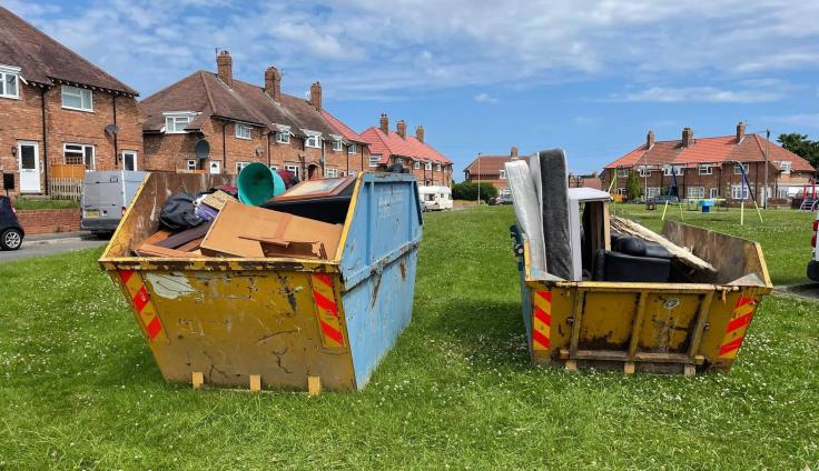Two large skips in a field in front of houses, filled with rubbish.