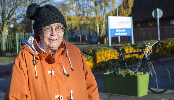 An elderly woman with glasses wears an orange raincoat coat and a black wool hat. Behind her is an open space with trees against a blue sky, a building, a wooden fence and a blue and white sign that reads "Birchwood Health Centre". There is also a topiary bush with yellow berries in it, and a blue bicycle attached to a green planter.