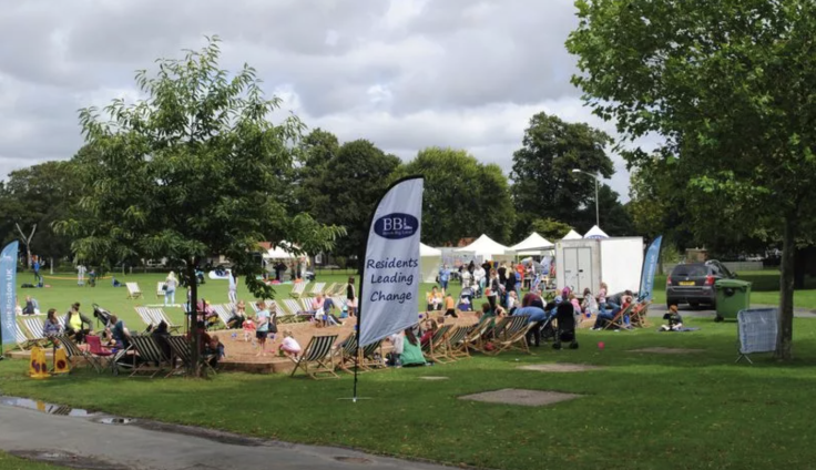 Boston Beach organised by Boston Big Local takes place in the local park. A large sand pit is surrounded by groups of people sitting in stripy deck chairs, white tents and a flag that reads 'BBL Residents Leading Change'.