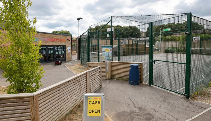 A sandwich board saying 'Cafe Open' in front of a single storey brick building and green steel-framed play area