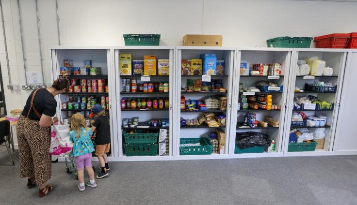 A row of glass-fronted fridges, stocked with food, with a woman and two children with shopping bags.