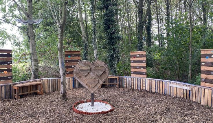 A woodland glade area, covered with bark and surrounded by trees, with pallet fences and a large wooden heart erected in the centre.