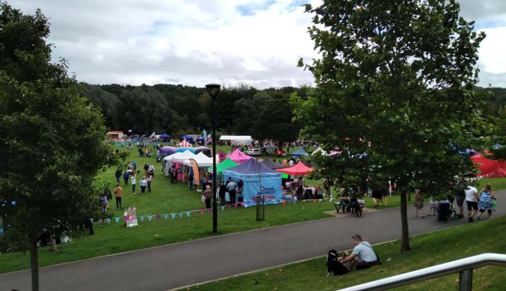 An open green area surrounded by trees, with colourful gazebos and crowds of people milling around.