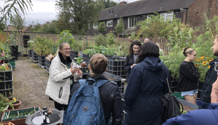 A group of volunteers, wearing waterproof jackets and coats, stand in a community garden. They are surrounded by multiple growing beds, filled with different plants. Most people are facing one woman who is speaking to the group.