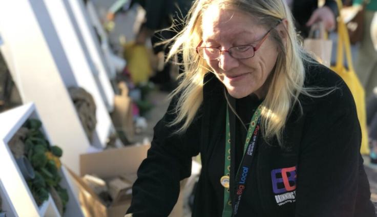 A woman with long blonde hair, glasses, wearing a black fleece and a green lanyard looks down at her hands out of frame.