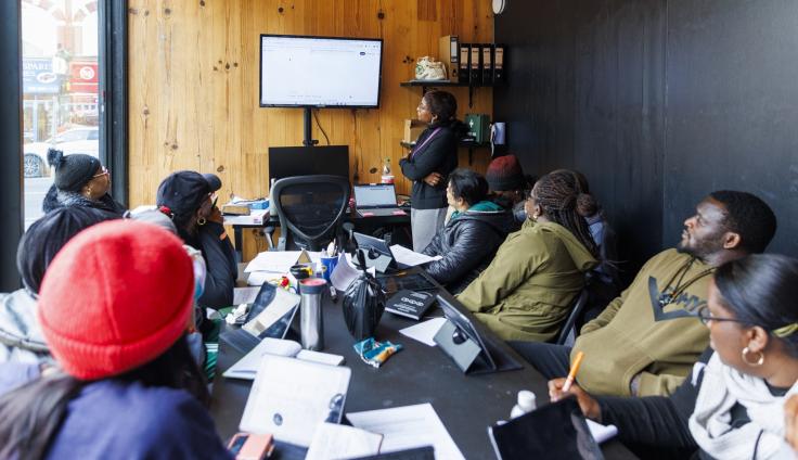 A group of people watching a woman who is facing a presentation screen at the front of the class.