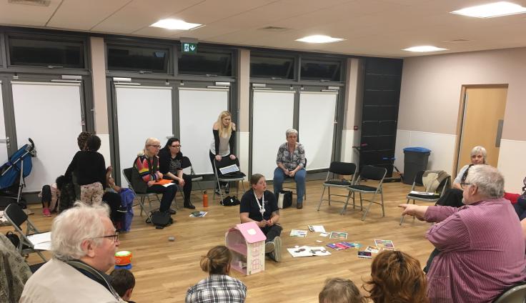 A multi-generational group gathers in a modern community room with large windows and wood floors. Adults and children sit in a loose circle on black folding chairs, with some participants sitting on the floor. A blonde woman in a black sleeveless top sits in the centre. Scattered on the wooden floor are books, papers, and a pink dollhouse.