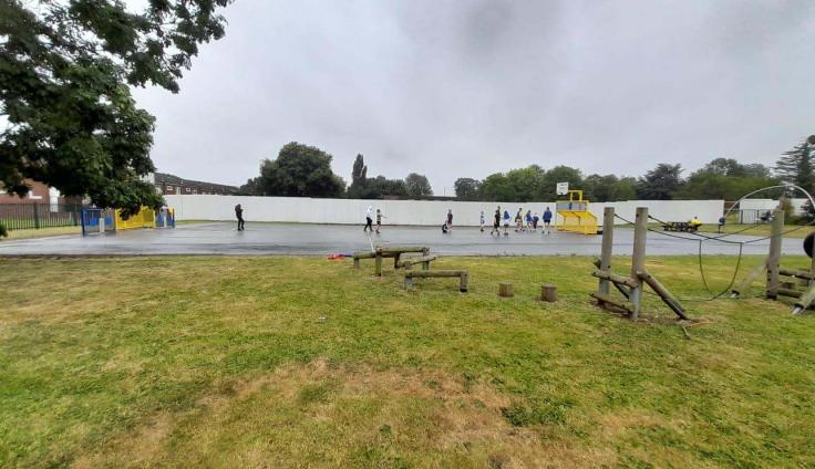 Children playing on a tarmac surface adjacent to a grassy area with wooden play equipment.